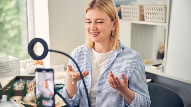 Excited bijouterie specialist presenting new necklace while keeping eyes at phone camera