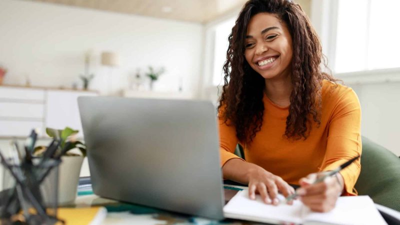 Distance Education. Portrait of smiling woman sitting at desk, using laptop and writing in notebook, taking notes, watching tutorial, lecture or webinar, studying online at home looking at screen